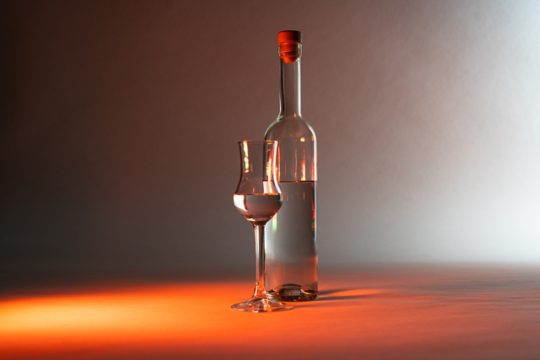 A kép közepén hosszú nyakú pálinkásüveg, tőle balra talpas pálinkáspohár van. Mindkettő félig tartalmaz pálinkát.