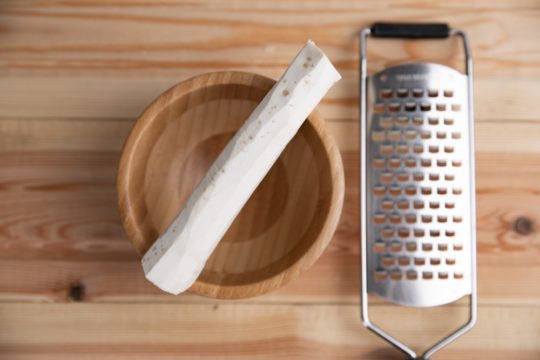 A képen középen barna színű fa kistálon egy meghámozott torma van, a jobb oldalán egy fém reszelő.