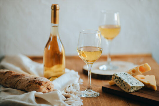 A kép jobb oldalán vágódeszkán különböző sajtok, közepén két pohár bor, jobb oldalán egy egész üveg bor és bagette.