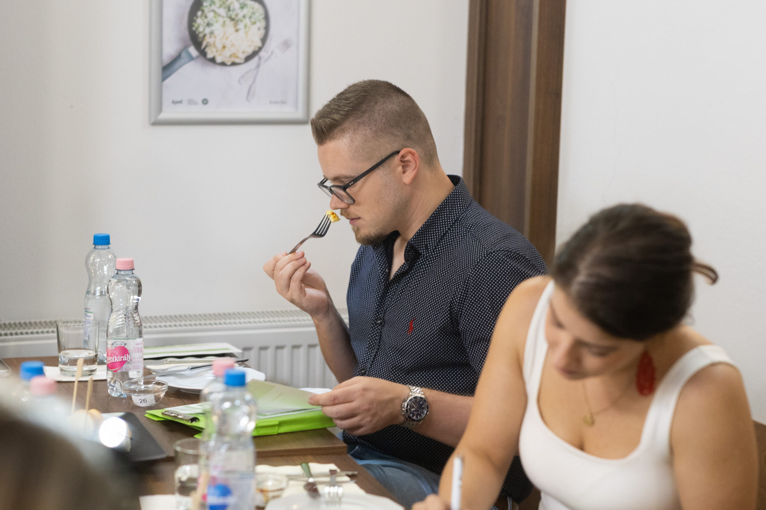 A képen egy női és egy férfi kóstoló van, a férfi a sajt-falatot vizsgálja.