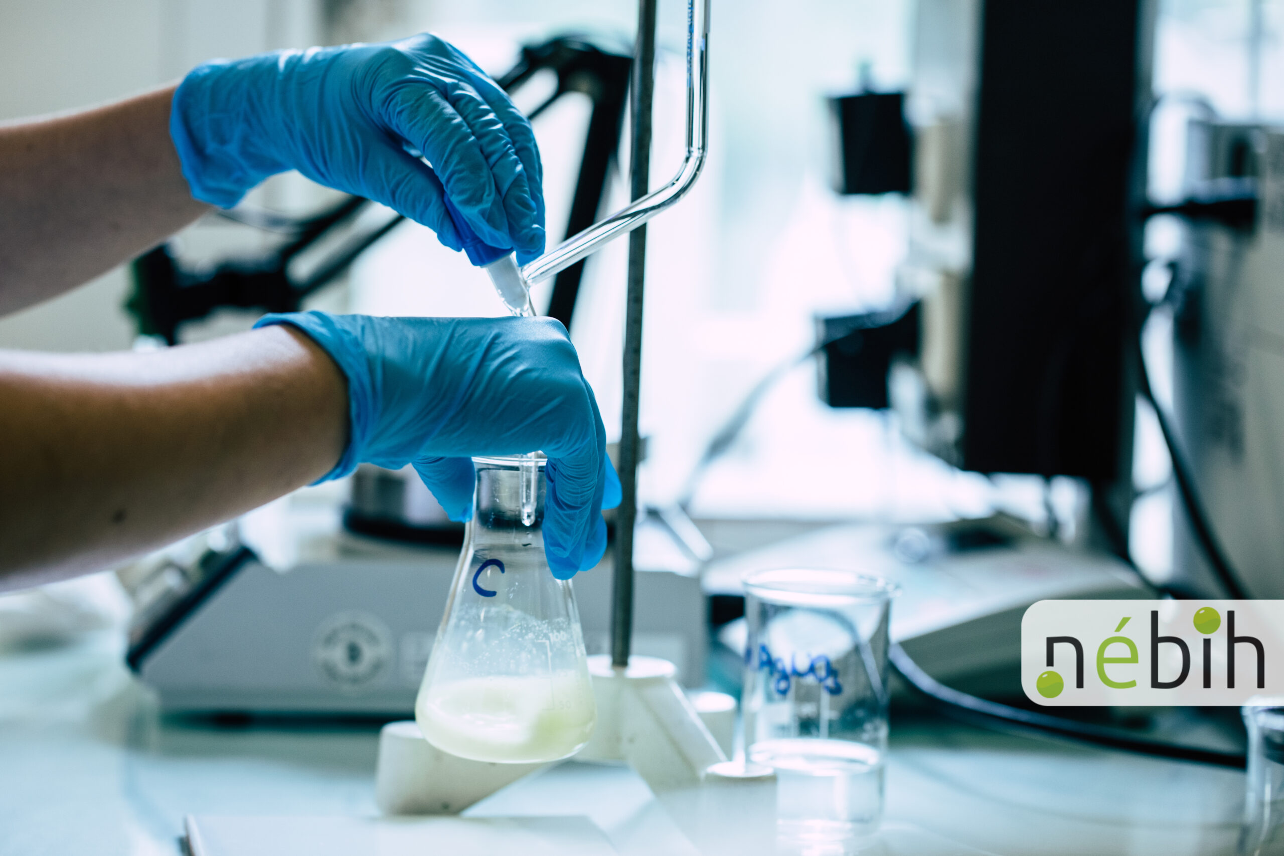 Laboratóriumi vizsgálat során védőkesztyűs kézzel folyadékot adagolnak egy lombikba.
